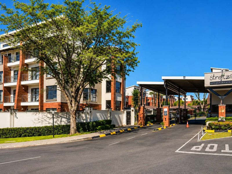 The entrance to Strelitzia Lifestyle Estate featuring a modern security gatehouse, paved driveway, and multi-story apartment buildings with brick and render finishes.