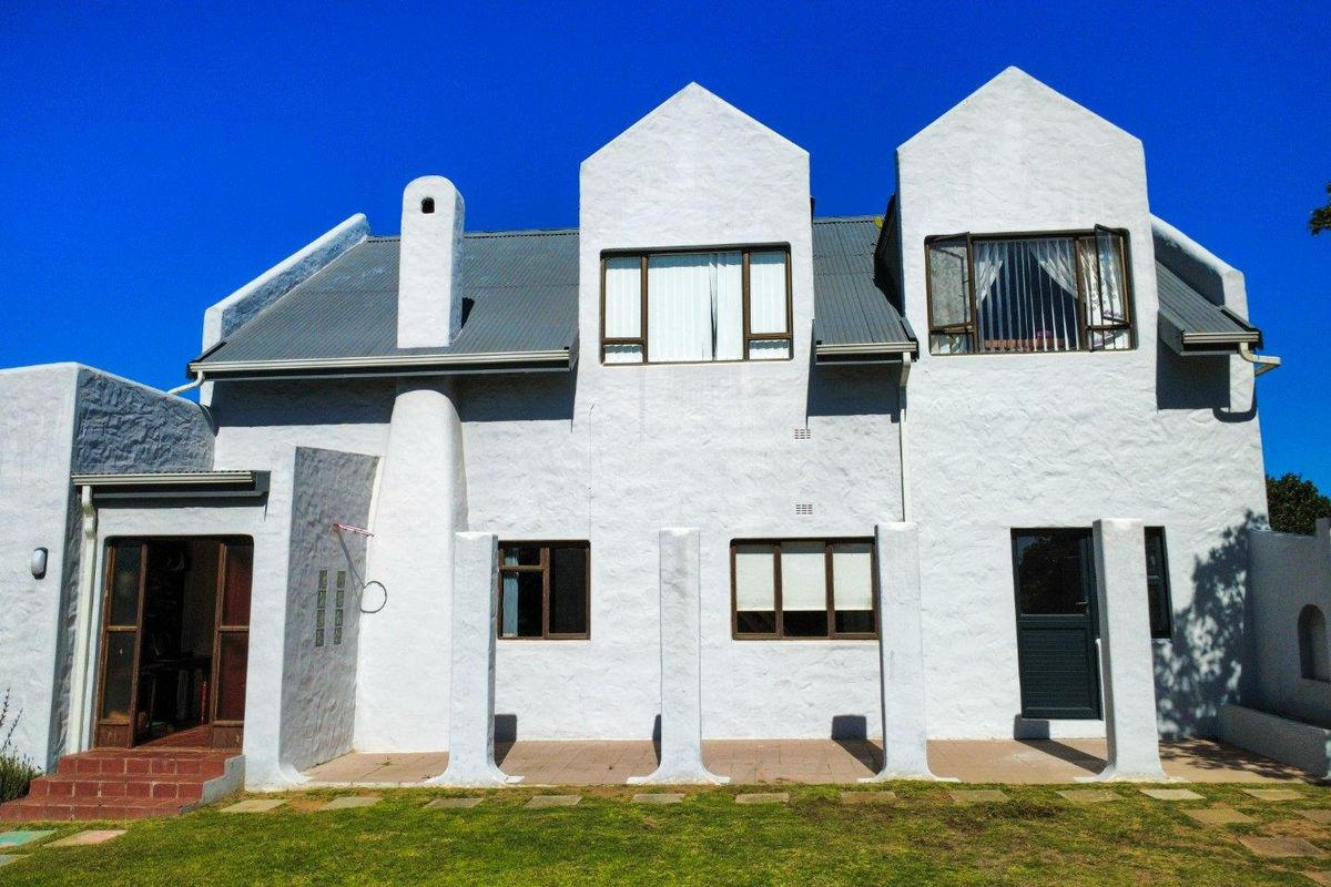Rear elevation of a multi-story house featuring a textured white facade, corrugated metal roof, chimney, and a paved patio area overlooking the garden.