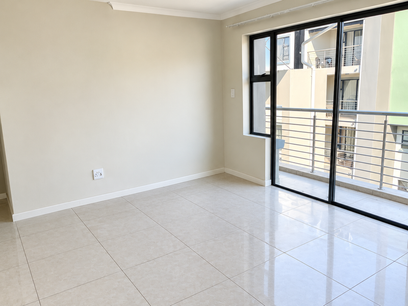 A bright, empty living area featuring neutral tiled flooring and a large sliding glass door providing access to a private balcony.