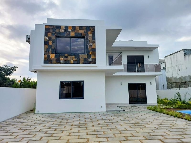Modern two-story residential exterior featuring clean white render, stone cladding accents, glass balcony railings, and paved outdoor area.