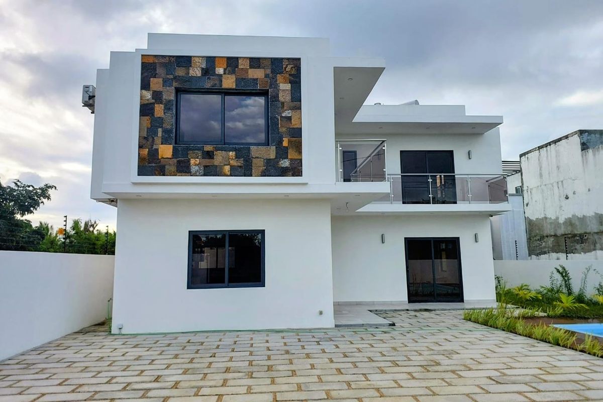 Modern two-story residential exterior featuring clean white render, stone cladding accents, glass balcony railings, and paved outdoor area.