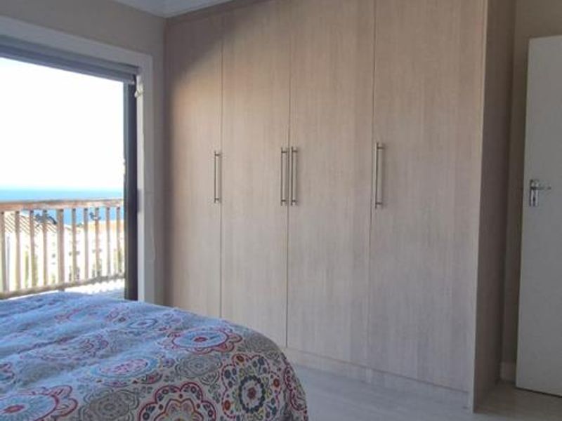 A bright bedroom featuring light-toned laminate flooring, ample built-in cupboards, and a sliding door providing access to a balcony with scenic views.