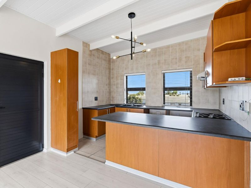A clean, functional kitchen featuring wooden cabinetry, dark laminate countertops, a built-in hob, and a bright, open layout with exposed beam ceilings.