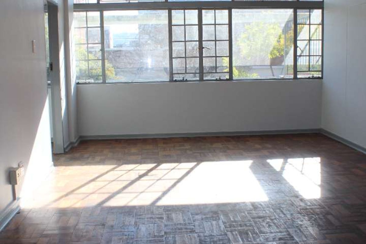 An empty living room featuring original parquet flooring with some missing tiles and a large window providing natural light.