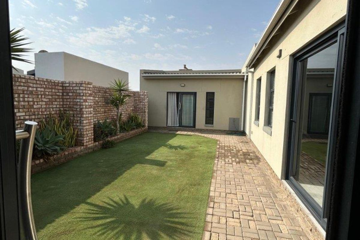 A well-maintained rear garden featuring a paved walkway, artificial lawn, and a brick boundary wall, accessible via sliding glass doors.