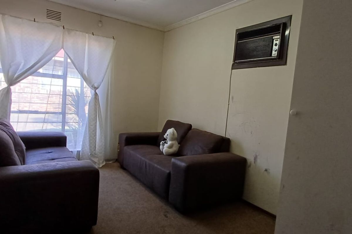 A living room featuring a wall-mounted air conditioning unit, built-in cupboard, and window security bars, requiring cosmetic updates to the walls and flooring.