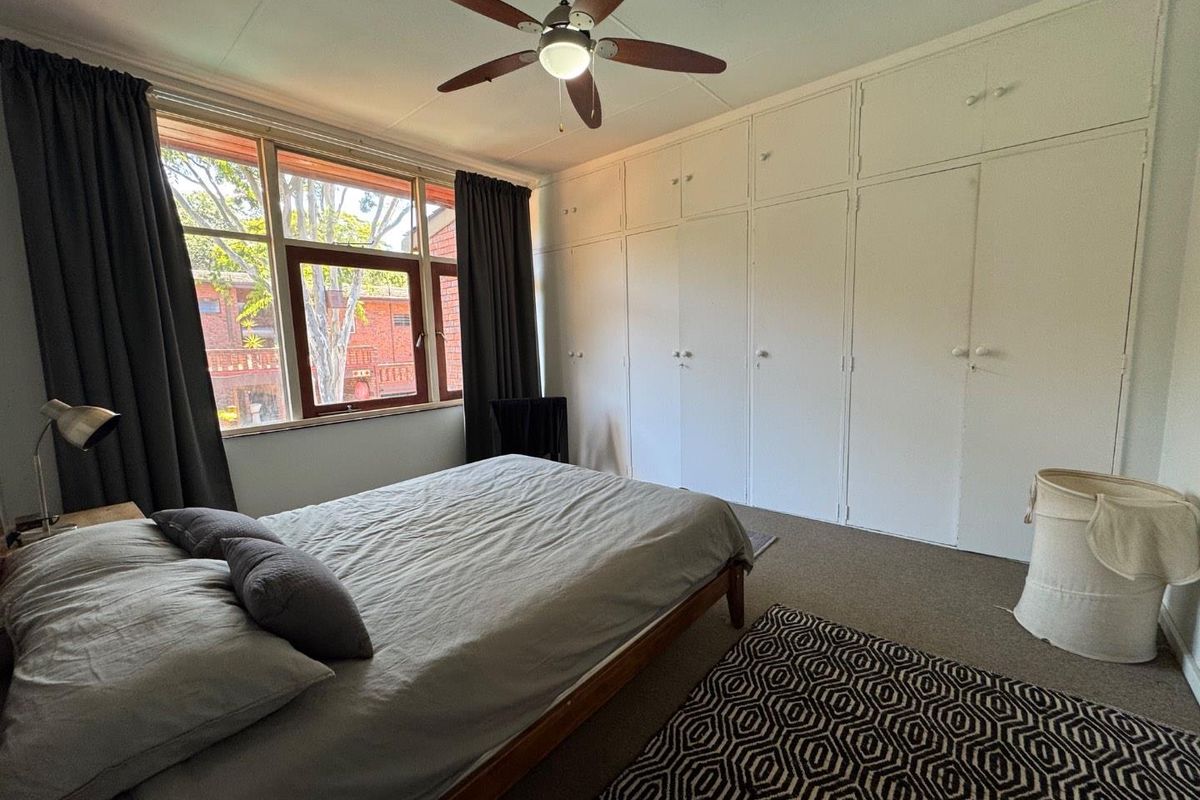 A bedroom featuring a large wall of built-in cupboards, a ceiling fan, and large windows providing natural light.
