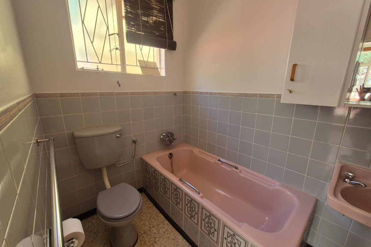 A dated bathroom featuring original pink sanitary ware, light blue wall tiling, and a window with security bars.