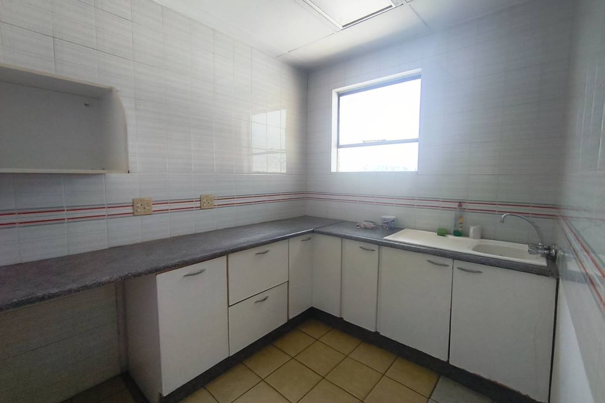 A functional, empty office kitchen featuring white built-in cabinetry, laminate countertops, a single sink, and tiled finishes throughout.