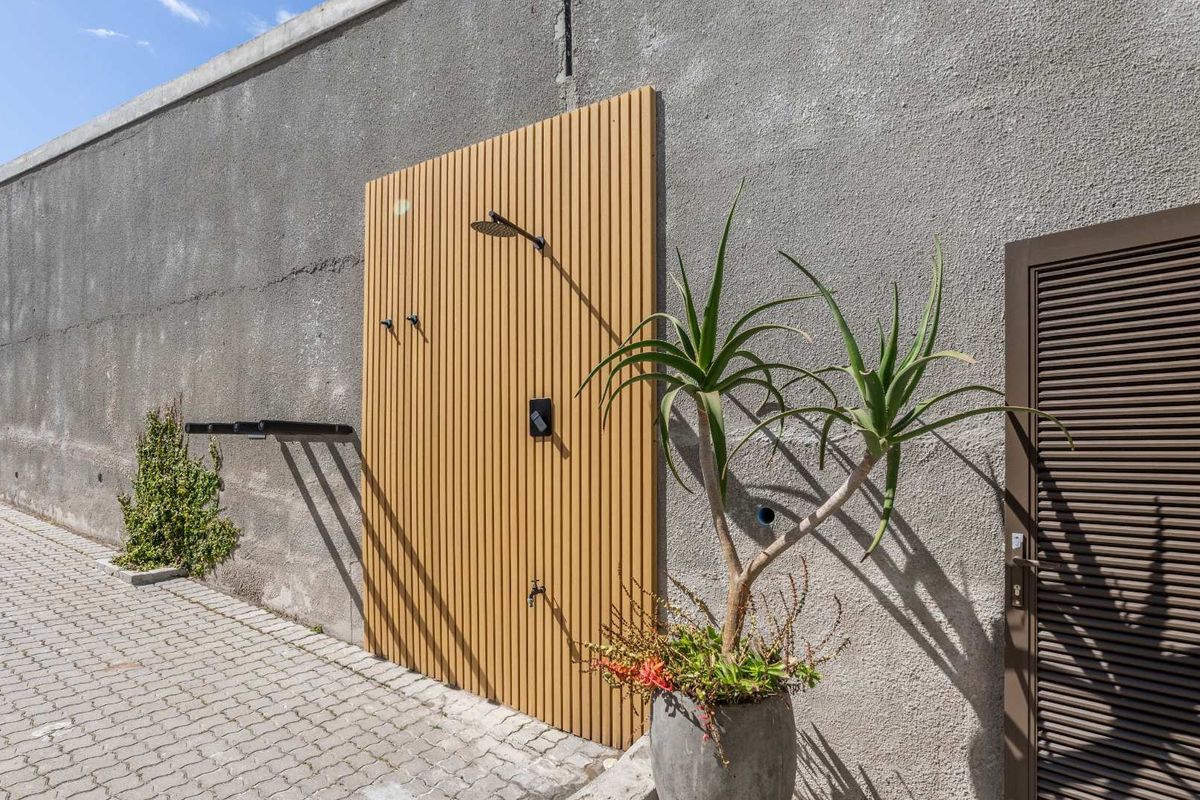 Outdoor shower area featuring a modern slatted timber panel, wall-mounted shower head, and tap, set against a textured concrete boundary wall.