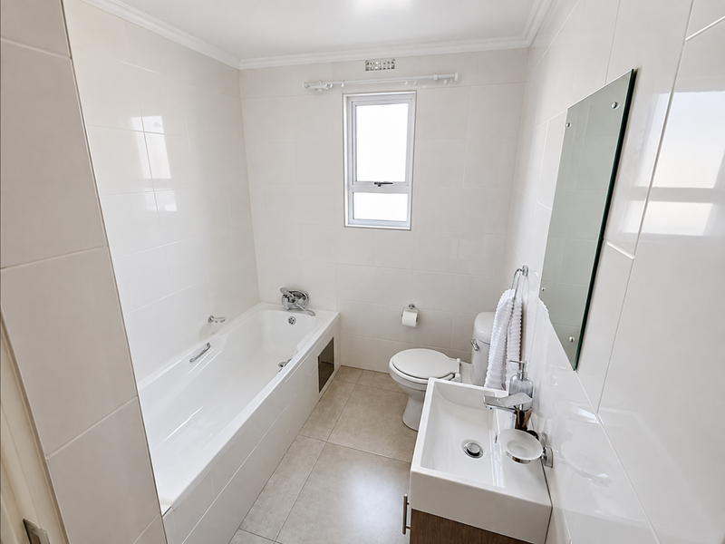 A clean, modern bathroom featuring a built-in bathtub, vanity basin, wall-mounted toilet, and neutral floor-to-ceiling wall tiles.