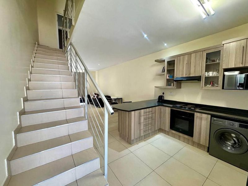 Open-plan kitchen featuring modern wood-finish cabinetry, integrated appliances, and tiled flooring, adjacent to a staircase with a metal balustrade.