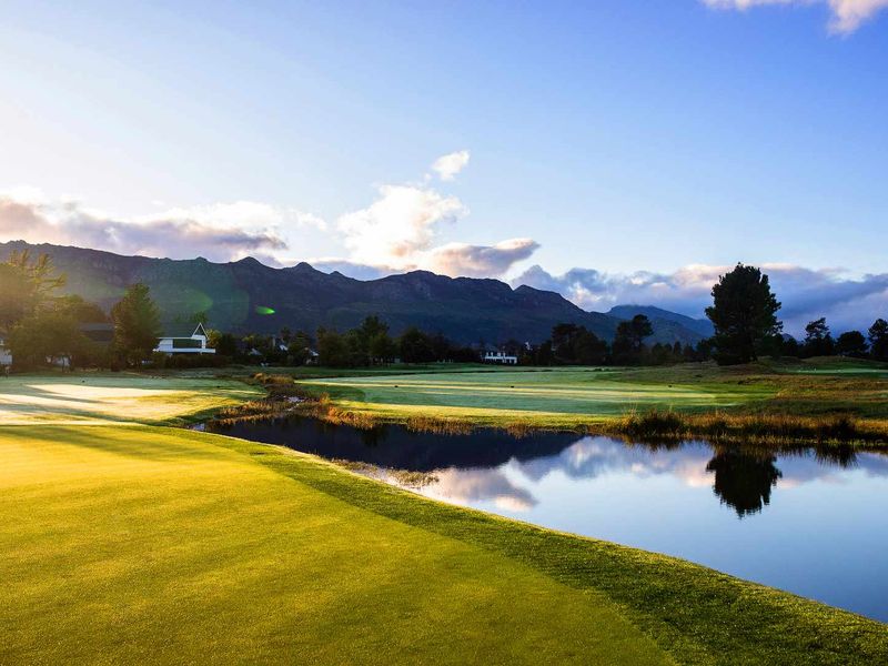 A scenic view of a well-maintained golf course featuring a tranquil water hazard with a mountain range backdrop under a clear sky.