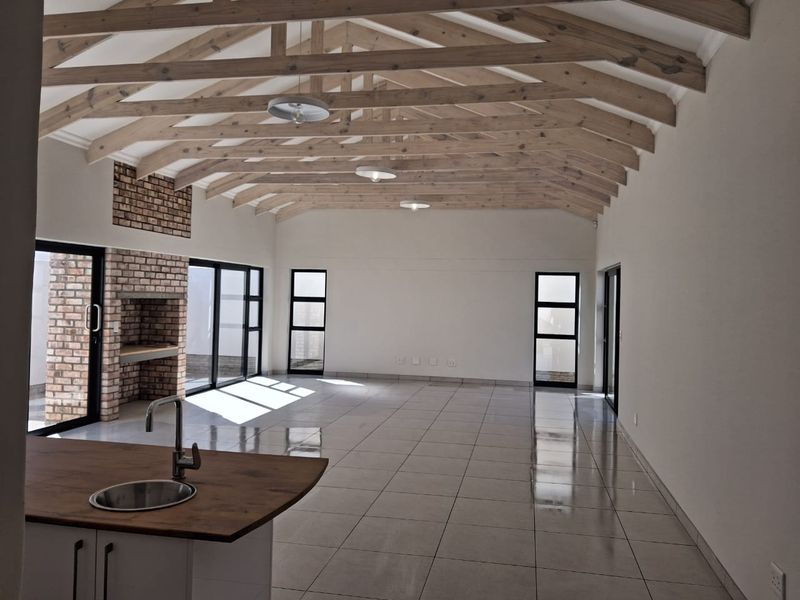 Spacious open-plan living area featuring high vaulted ceilings with exposed timber trusses, tiled floors, a brick fireplace, and direct access to the outdoor area.