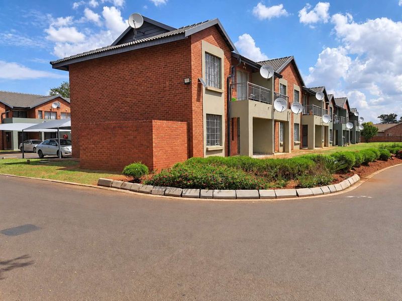 A view of a multi-unit apartment complex featuring face brick construction, tiled roofs, private balconies, and established garden beds with paved driveway access.
