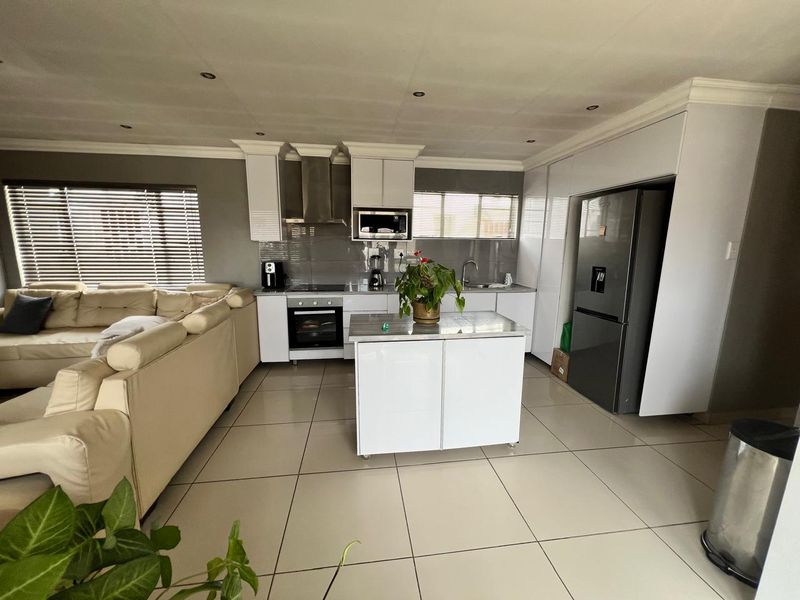Modern open-plan kitchen and living area featuring sleek white cabinetry, built-in appliances, a central island, and neutral tiled flooring throughout.