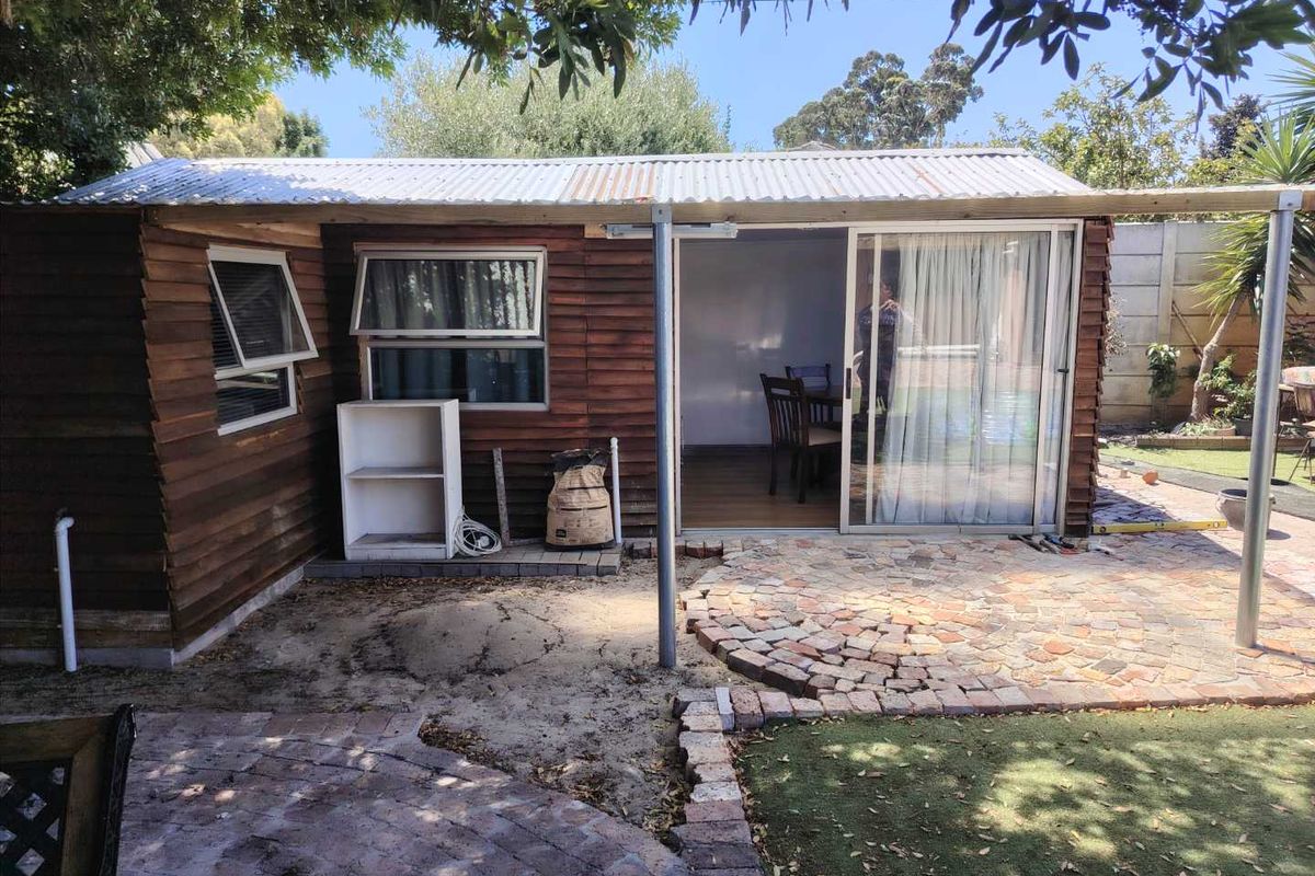 Rear view of a timber-clad cottage featuring a corrugated iron roof, sliding glass doors, and a brick-paved patio area in a garden setting.