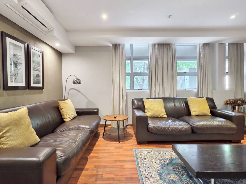 A well-maintained living room featuring timber flooring, recessed downlighting, and a split-system air conditioning unit, with large windows providing natural light.
