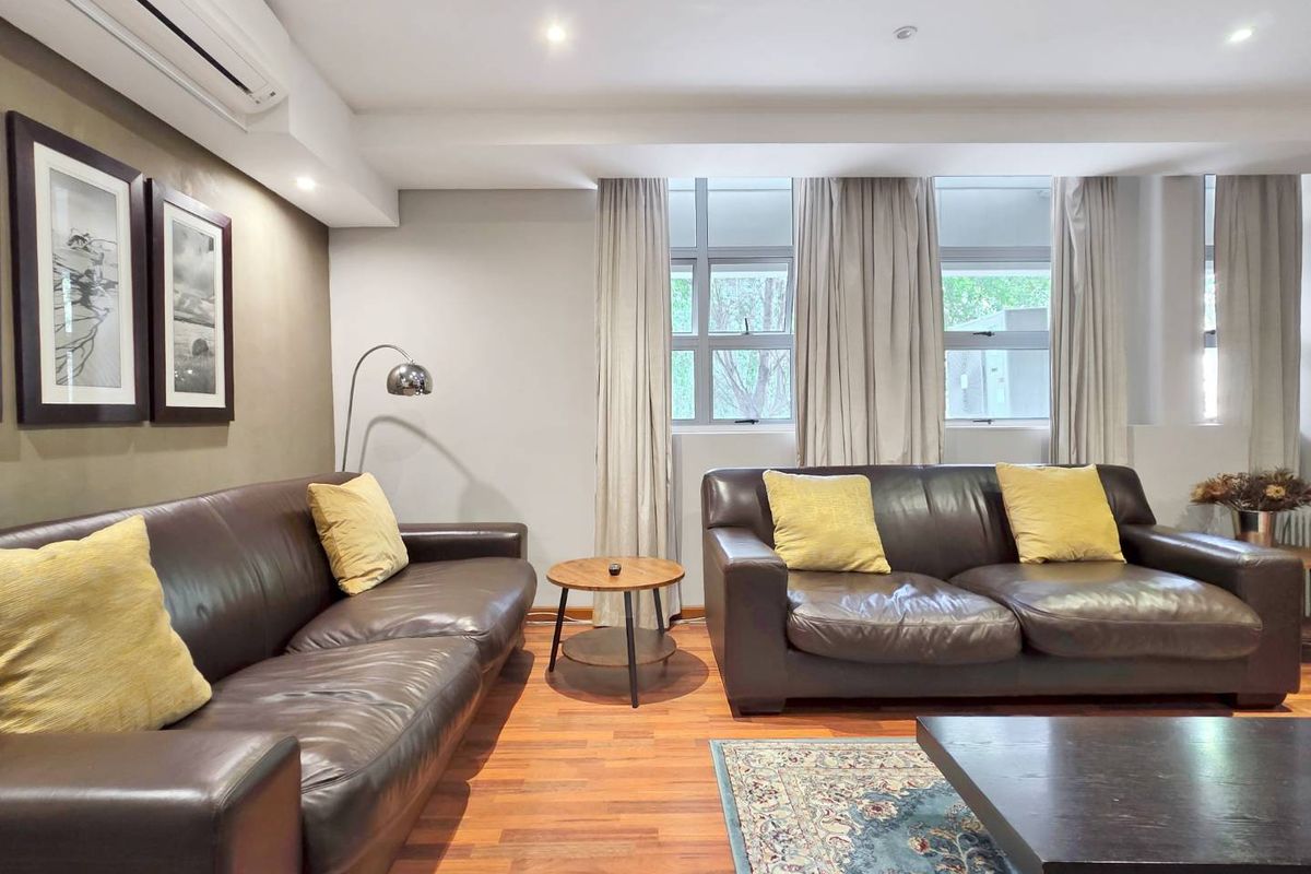 A well-maintained living room featuring timber flooring, recessed downlighting, and a split-system air conditioning unit, with large windows providing natural light.