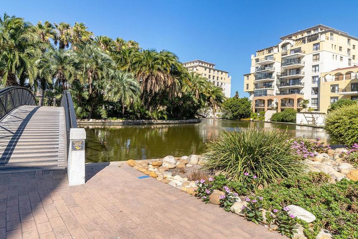 A scenic view of a pedestrian bridge leading over a tranquil pond, surrounded by lush landscaping and apartment complex architecture.