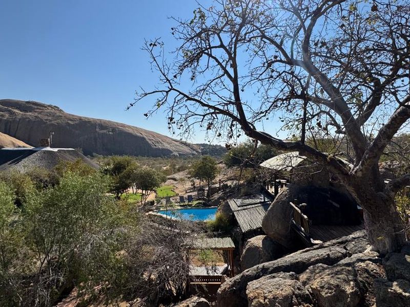 Scenic elevated view of the lodge grounds featuring a swimming pool, natural rock formations, and traditional thatched roof structures set in a rugged landscape.