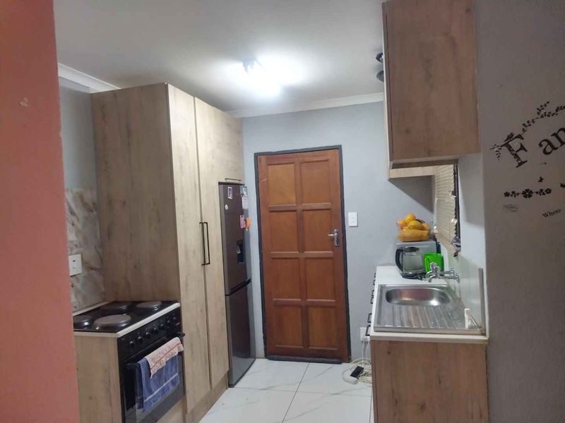 A compact kitchen featuring light wood-finish cabinetry, a stainless steel sink, and a freestanding stove with tiled flooring.