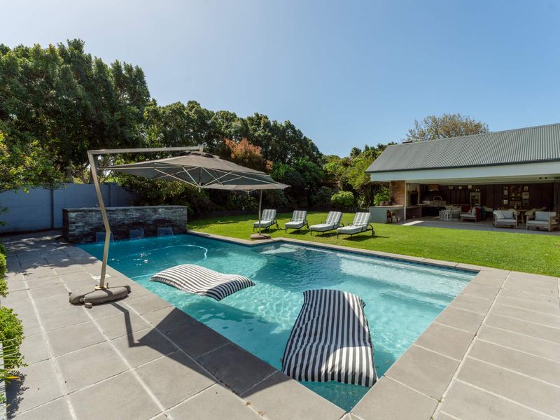 A well-maintained outdoor pool area featuring a stone water feature, paved decking, and a covered patio with a built-in braai.