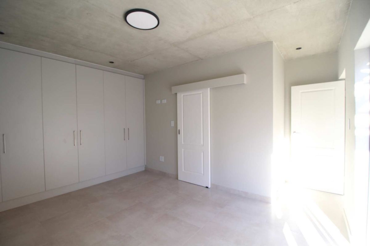 A bright, modern bedroom featuring neutral tiled floors, built-in cupboards, and an exposed concrete ceiling with contemporary lighting.