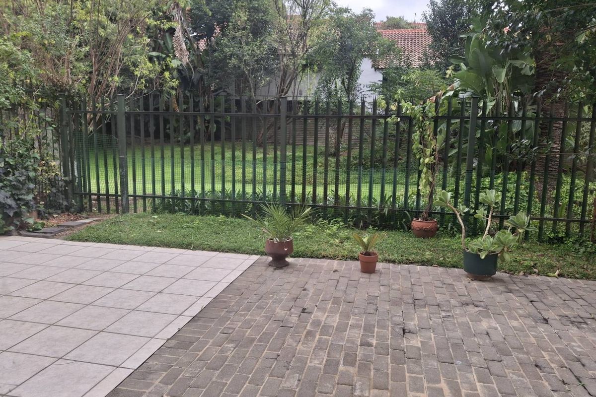 A rear outdoor area featuring a paved patio, lawn, and metal security fencing, providing a private garden space.