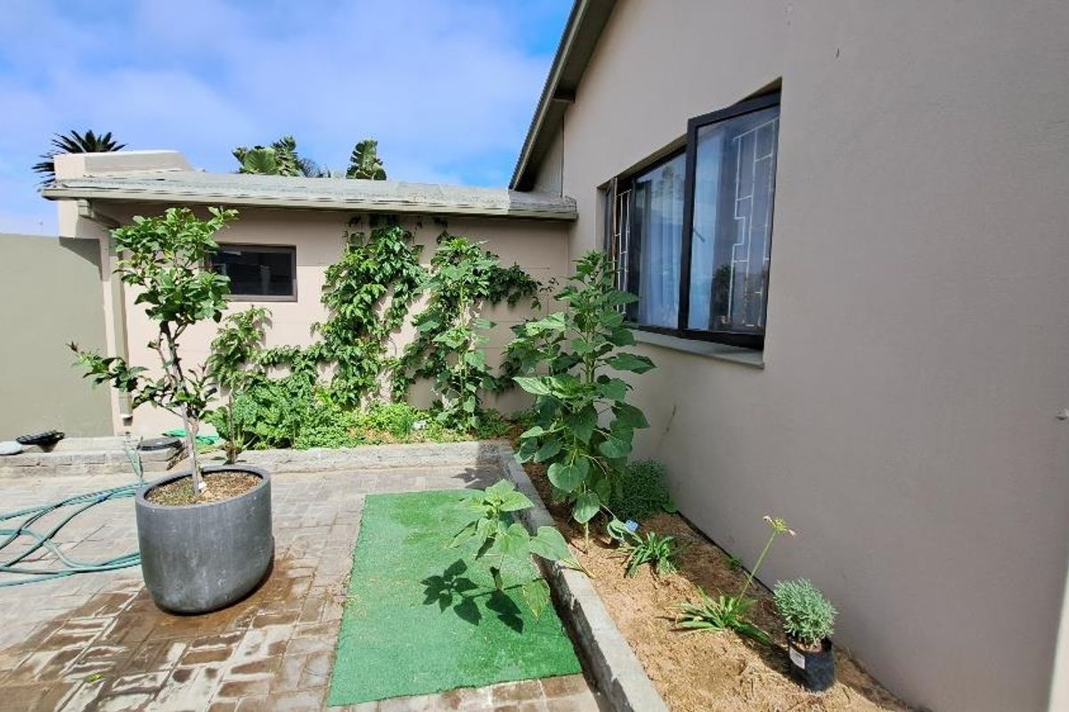 A rear exterior view of the property featuring a paved patio area, garden beds with established greenery, and a sliding window with security bars.