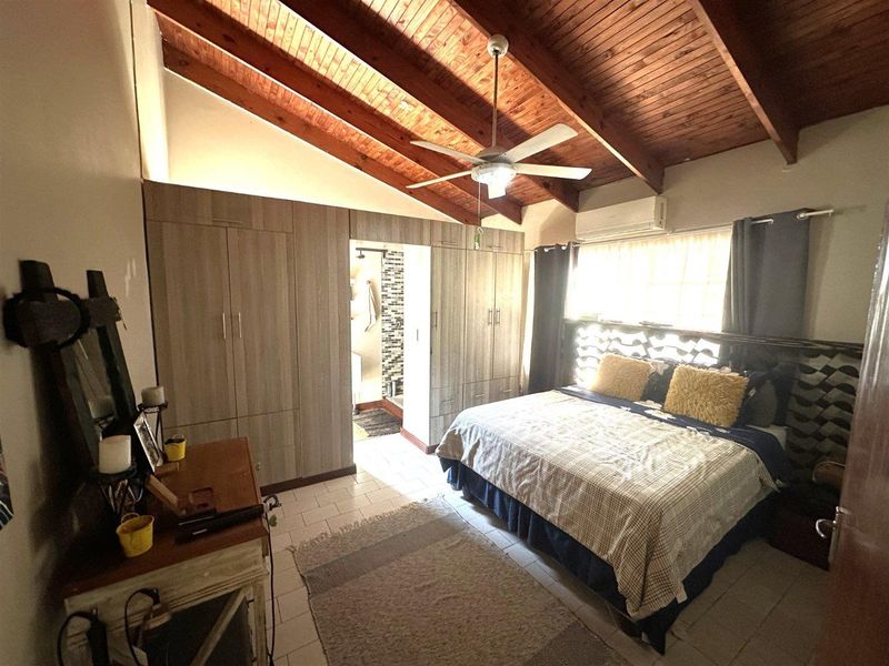 Spacious bedroom featuring exposed timber ceiling beams, built-in wardrobes, a ceiling fan, and an integrated air conditioning unit with direct access to an en-suite bathroom.
