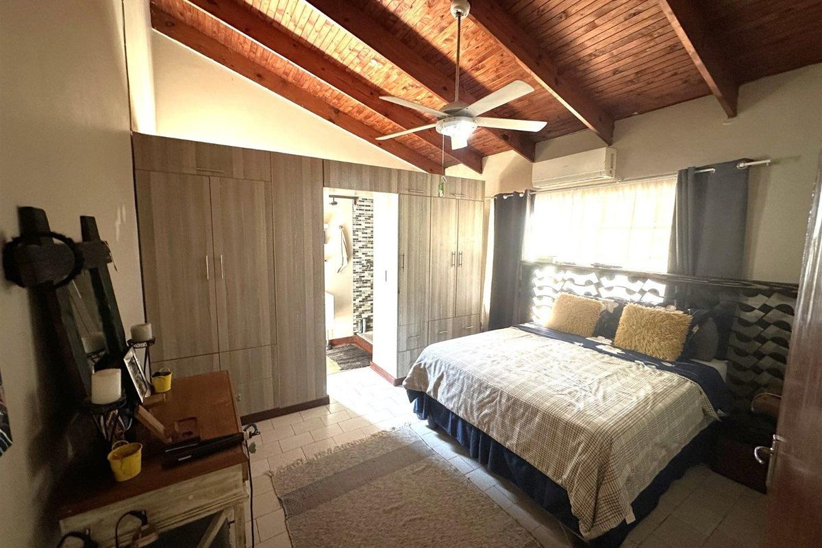 Spacious bedroom featuring exposed timber ceiling beams, built-in wardrobes, a ceiling fan, and an integrated air conditioning unit with direct access to an en-suite bathroom.