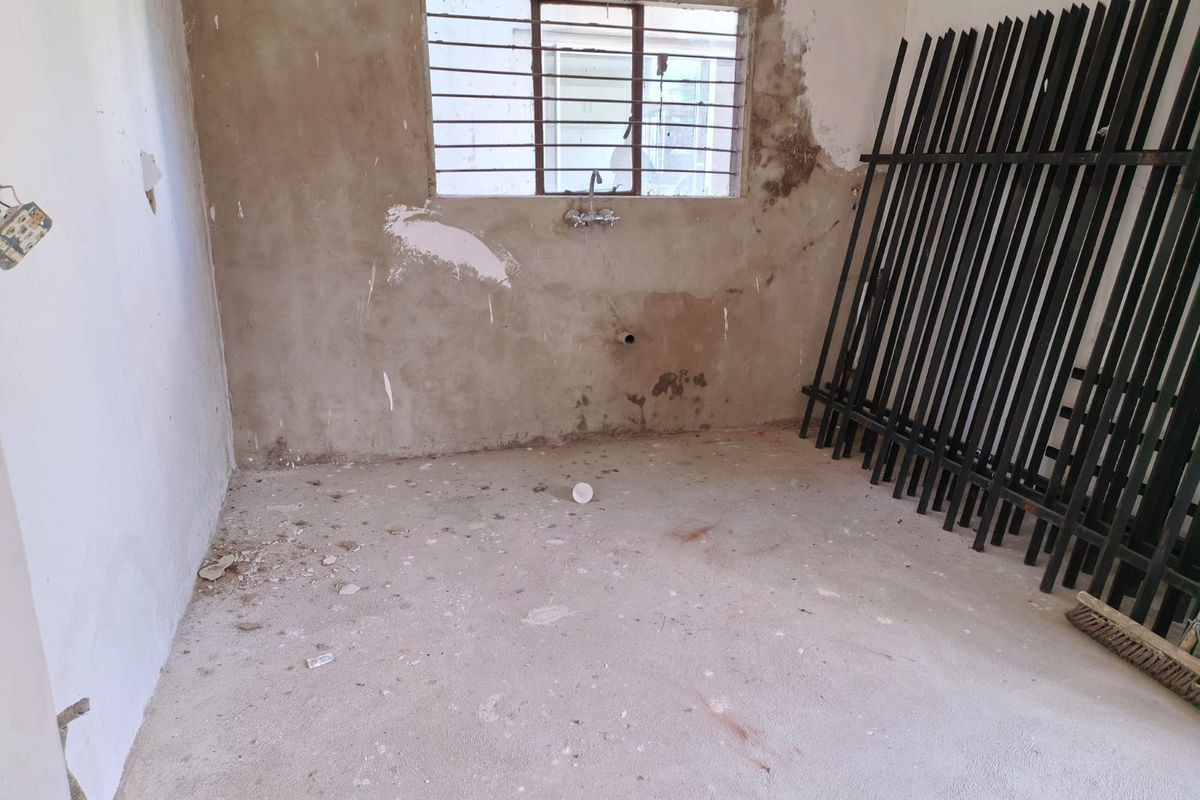 An unfinished kitchen space featuring exposed concrete walls, a wall-mounted faucet, and a window with security bars, requiring significant renovation and finishing.