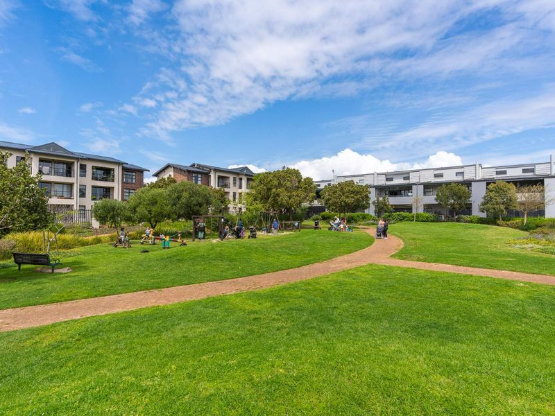 A well-maintained communal garden area featuring expansive green lawns, paved walking paths, and a playground, situated within an apartment complex.