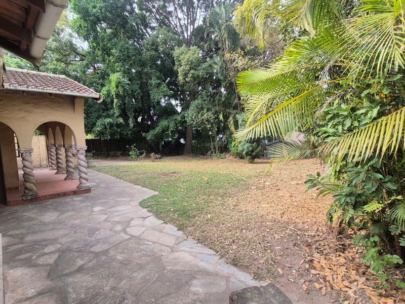 A rear garden view featuring a covered patio with decorative arched columns and stone paving, overlooking a spacious lawn area with mature trees.