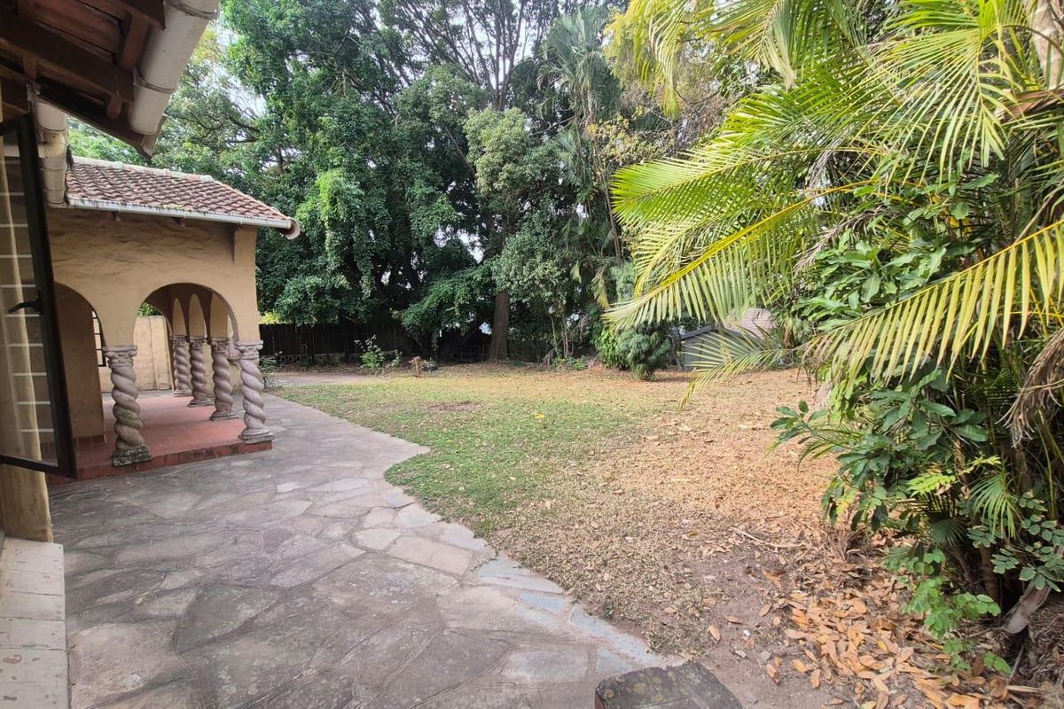 A rear garden view featuring a covered patio with decorative arched columns and stone paving, overlooking a spacious lawn area with mature trees.