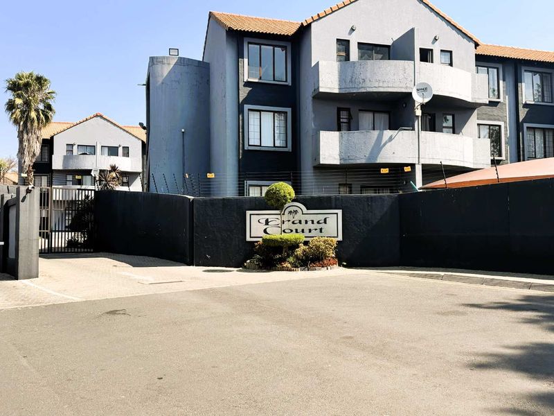 Front entrance view of Grand Court apartment complex featuring paved driveway, security gate, electric fencing, and multi-story residential building with balconies.