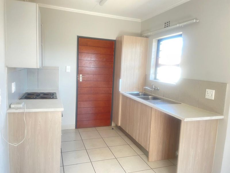 A functional kitchen featuring light wood-look cabinetry, laminate countertops, an electric hob, and a double sink with tiled splashbacks and flooring.