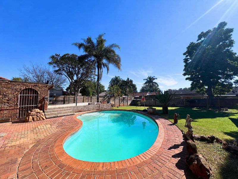 A backyard pool area featuring a kidney-shaped swimming pool surrounded by brick paving, a stone retaining wall, and a landscaped garden.