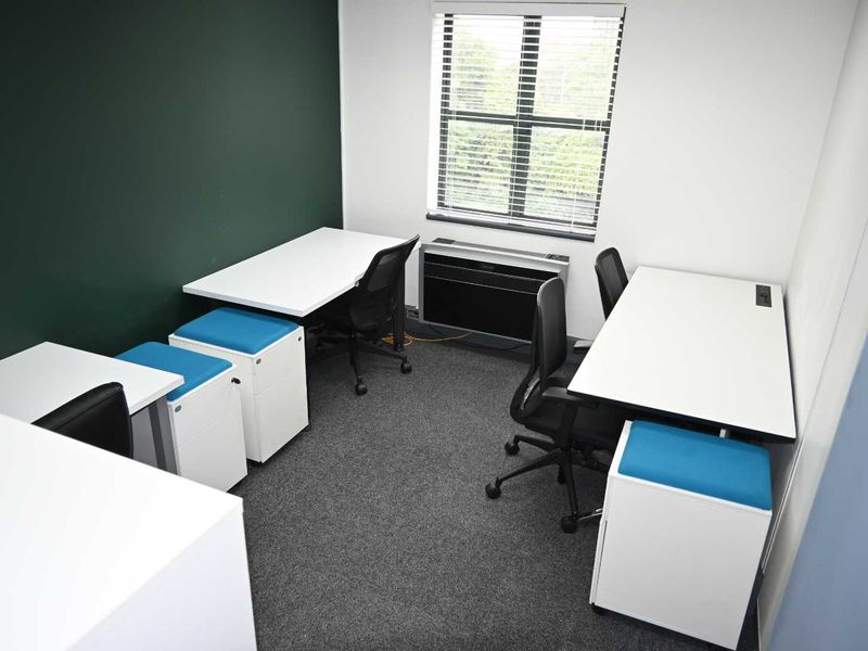 A functional office space featuring dark green accent wall, carpeted flooring, window blinds, and a built-in HVAC unit.