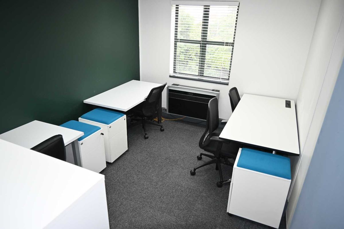 A functional office space featuring dark green accent wall, carpeted flooring, window blinds, and a built-in HVAC unit.