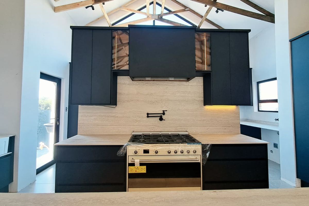 Modern kitchen featuring dark cabinetry, stone backsplash, gas stove with pot filler, and striking exposed timber roof trusses in a high-ceiling space.