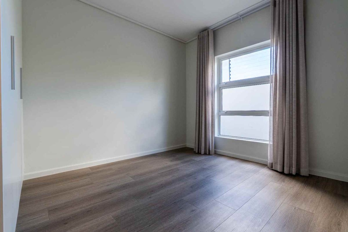 A bright, empty bedroom featuring modern laminate flooring, built-in cupboards, and large windows with neutral curtains.