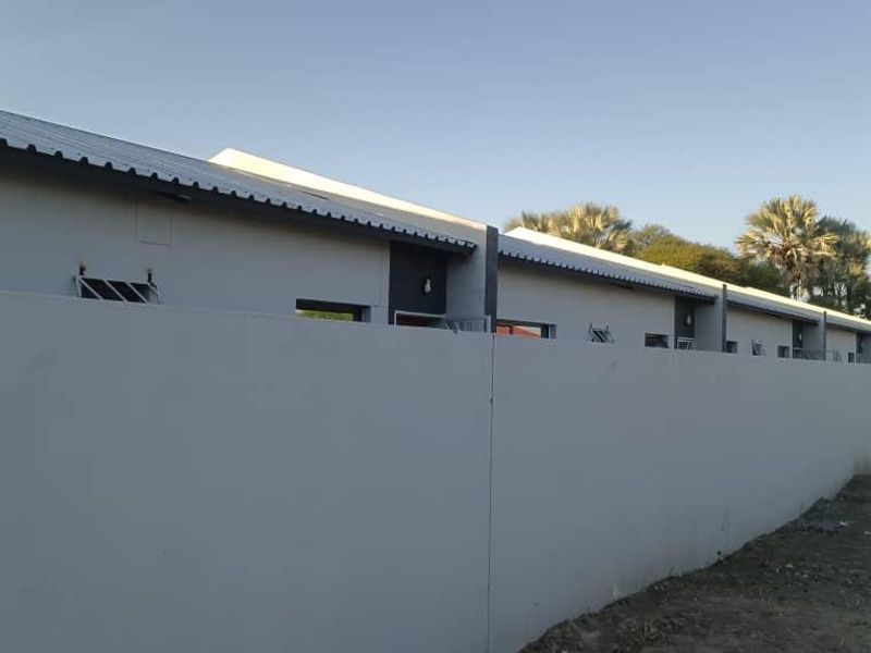 A view of a row of residential units featuring a long perimeter boundary wall, corrugated metal roofing, and security bars on the windows.