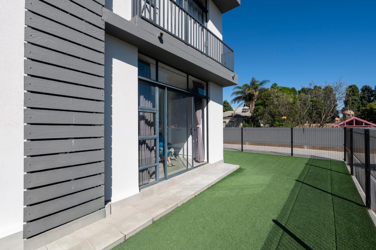 Rear view of the apartment building featuring a paved patio area, artificial turf lawn, sliding glass doors, and an upper-level balcony with metal railings.