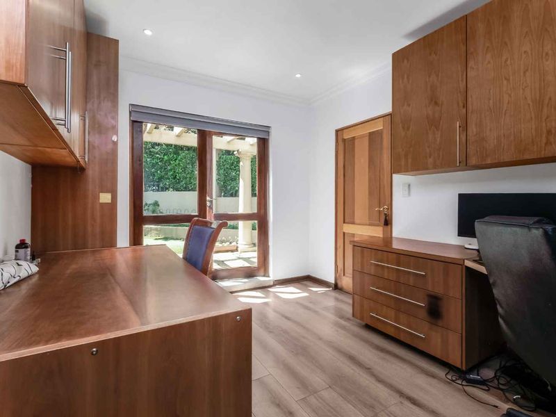 A well-appointed home office featuring extensive built-in wooden cabinetry, a spacious desk, laminate flooring, and direct access to the garden through a glass door.