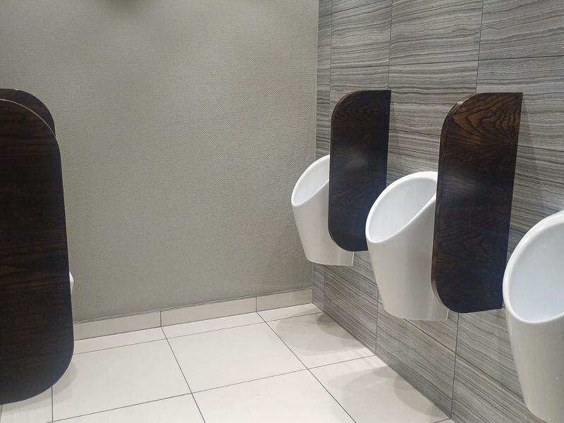 A clean, modern office restroom featuring wall-mounted urinals with dark wood privacy partitions and contemporary grey tiled wall finishes.