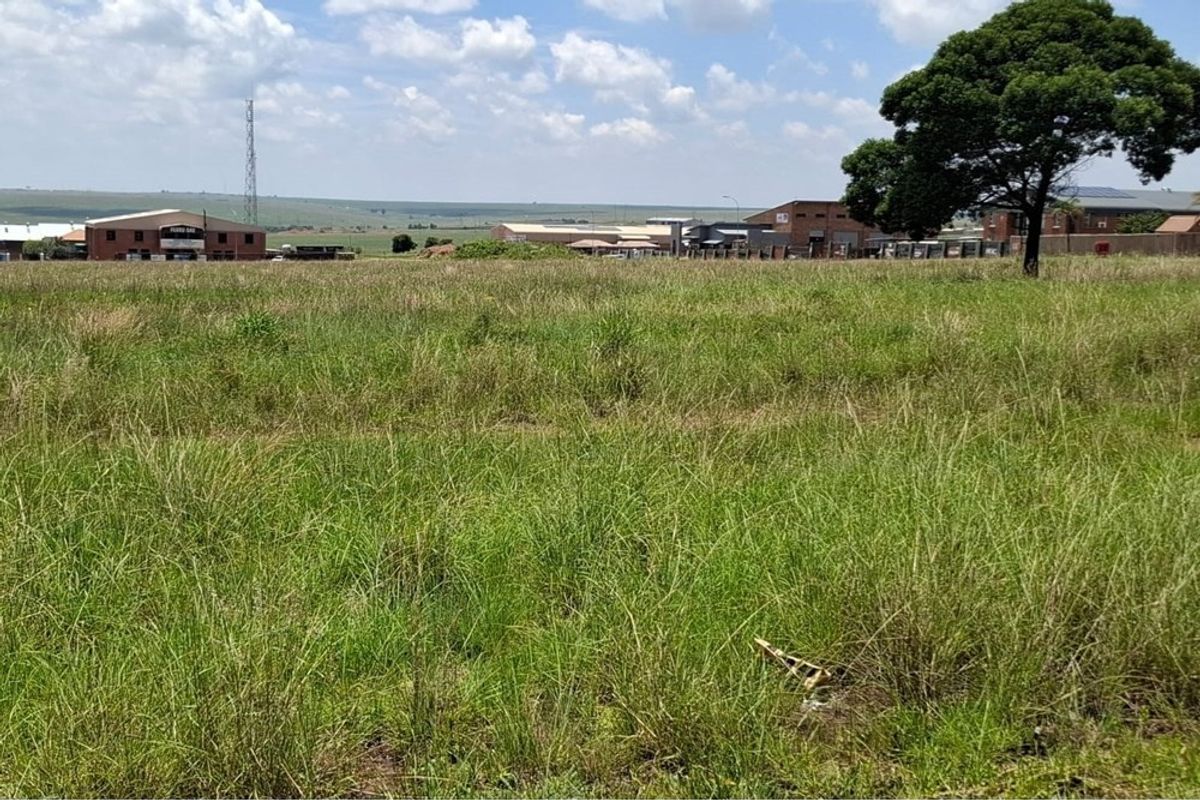 A view of a vacant residential plot featuring natural grass cover and a mature tree, with surrounding commercial and industrial buildings in the distance.