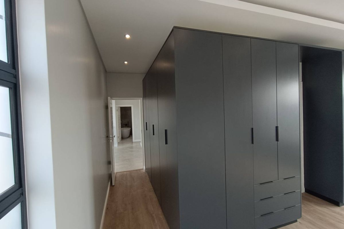 Modern bedroom featuring extensive dark grey built-in cabinetry, recessed ceiling lighting, and light wood-look laminate flooring with a view into an adjacent bathroom.