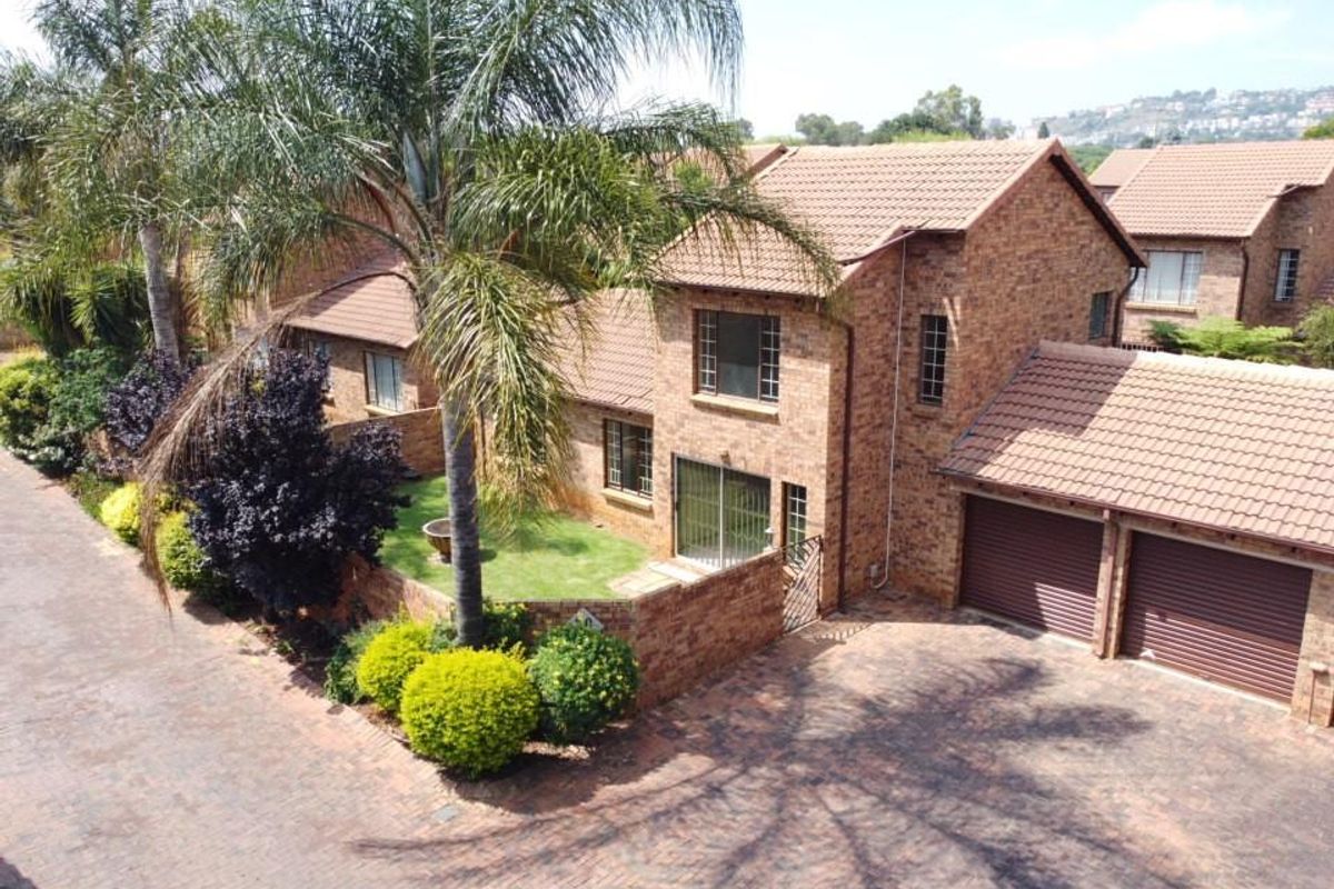 Aerial view of a brick townhouse featuring a double garage, paved driveway, and a well-maintained private garden with mature landscaping.